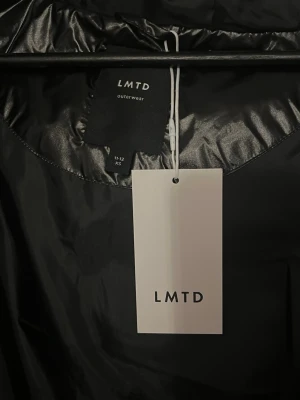 Svart pufferjacka från LMTD - Snygg svart pufferjacka från LMTD med glansig finish och huva. Jackan har en rak passform och dragkedja framtill. Perfekt för kalla vinterdagar och enkel att matcha med olika outfits.