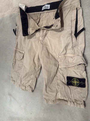 Beige cargoshorts från Stone Island - Säljer ett par beige cargoshorts från Stone Island Junior med flera fickor och den klassiska logotypen på benet. Shortsen har elastiska benslut och detaljer i svart vid midjan. Perfekta för sommaren och riktigt snygga till en avslappnad stil. JUNIOR STORLEK 164/158