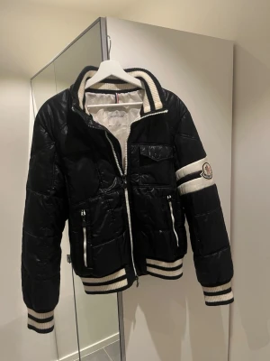 Svart pufferjacka från Moncler - Tja, säljer en svart pufferjacka från Moncler med vita detaljer och ribbade muddar. Jackan har dragkedja framtill, fickor med dragkedja och en vit Moncler logga på ärmen.