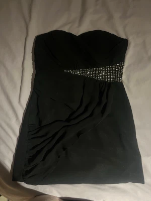 Svart tubtopp/kort klänning med strass - Snygg svart axelbandslös tubtopp/kort klänning med glittrande pärldetaljer framtill och draperad effekt. Toppen har en elegant och figurnära passform, perfekt för att sticka ut. Materialet känns mjukt och följsamt.