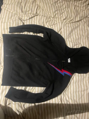 Bvaldi kashmir hoodie - Säljer en svart hoodie med dragkedja och huva. På bröstet finns en snygg blixtdetalj i rött och blått som ger en cool kontrast. Hoodien har långa ärmar och är tillverkad i mjukt material, perfekt för en avslappnad stil.