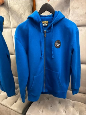 Blå hoodie från Napapijri - Blå hoodie med dragkedja från Napapijri. Har huva med snörning, två fickor fram och ett runt märke på bröstet. På baksidan av huvan finns en gul logotyp. Storleken L men kan passa M.