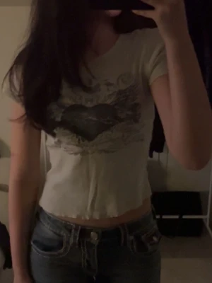 Vit croppad topp med tryck - Trendig vit croppad topp med korta ärmar och ett stort hjärtformat tryck framtill. T-shirten har rå kant nertill för en avslappnad vibe och är perfekt att styla med jeans. Passformen är slim och materialet känns mjukt mot huden.