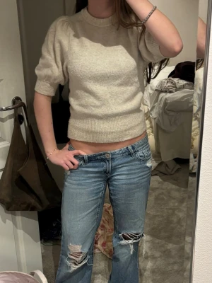 Beige puffärm stickad tröja - Säljer en beige stickad tröja med puffärmar och ribbade muddar. Tröjan har en croppad passform och rund halsringning. Perfekt för dig som gillar en trendig och mysig stil. Materialet känns mjukt och bekvämt.