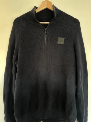 Svart stickad half zip tröja från BOSS - Snygg svart stickad tröja från BOSS med halv dragkedja och ribbstickad struktur. Storlek L.  Tröjan har en diskret BOSS-logga på bröstet och hög krage som ger en stilren look. Perfekt för dig som gillar klassisk och enkel design.