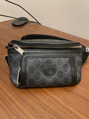 Svart axelväska från Gucci - Snygg svart axelväska från Gucci med klassiskt GG-mönster i grått. Väskan har två dragkedjefack och justerbar axelrem i textil. Perfekt för att bära det viktigaste och ger en lyxig touch till din outfit.