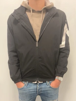 Svart Moncler vindjacka med huva - Svart vindjacka från Moncler med vit M-detalj på ärmen och logotyp på axeln. Jackan har dragkedja framtill, två sidofickor och elastiska muddar vid ärmslut och midja. Perfekt för en sportig och stilren look.