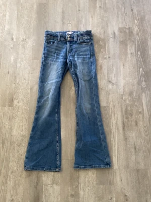 Blå bootcut jeans - Säljer ett par klassiska blå bootcut jeans från Gina Tricot med snygga slitningar och broderade bakfickor. Jeansen har normal midja, fem fickor och stängs med dragkedja och knapp. Perfekta för en avslappnad och trendig look.