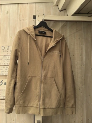 Beige hoodie från Peak Performance - Snygg beige hoodie från Peak Performance med dragkedja framtill, två stora fickor och broderad logga på bröstet. Tillverkad i mjukt material med borstad insida och justerbar huva med snören. Perfekt för chill dagar eller när du vill ha en clean look. HELT NY aldrig använd.  Pris kan diskuteras