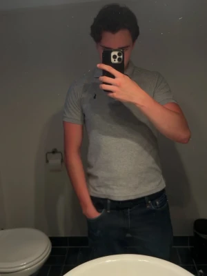 Grå pikétröja från Ralph Lauren - Klassisk grå pikétröja från Ralph Lauren med korta ärmar och svart broderad logga på bröstet. Tröjan har krage med knappar och är tillverkad i mjuk bomull. Perfekt för sommaren, Storlek M men sitter som S