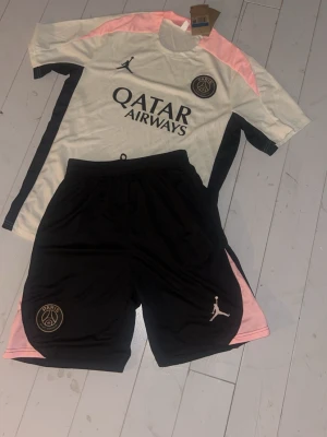 PSG träningsset Jordan x Qatar Airways - Säljer ett PSG träningsset med tröja och shorts i vitt, svart och rosa. Tröjan har Paris Saint-Germain och Qatar Airways-logga samt Jordan-märket. Shortsen är svarta med rosa detaljer och PSG-emblem. Perfekt för fotboll eller träning.
