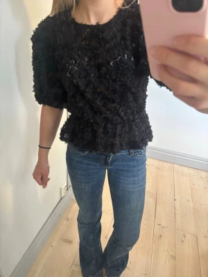 Svart blommig blus från Gina tricot - Supersnygg svart blus från Gina tricot med 3D-blommor över hela tyget. Blusen har korta puffärmar och rund halsringning. Materialet är polyester och ger en cool struktur. Perfekt för dig som vill sticka ut med en unik topp!