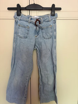 Ljusblå jeans med bälte från Zara - Ljusblå jeans från Zara med raka ben och två framfickor. Jeansen har ett snyggt denim-bälte med träfärgad rund spänne. Materialet är klassisk denim och modellen är avslappnad med hög midja. Perfekt för dig som gillar en cool och enkel stil.