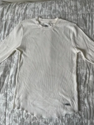 Vit ribbad långärmad topp från Corteiz - Vit långärmad topp från Corteiz med ribbad struktur och rund halsringning. Diskret logotyp på nederkanten. Perfekt för att skapa en clean och stilren look. Materialet är mjukt och bekvämt, vilket gör den enkel att bära hela dagen.