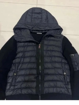Svart cardigan med huva från Moncler - Snygg svart dunväst från Moncler med stickade svarta ärmar och stor vadderad huva. Västen har dragkedja framtill, två fickor med tryckknappar och en Moncler-logga på ärmen. Perfekt för kyliga dagar och riktigt trendig look.