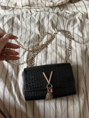 Svart axelväska med kedja från Valentino - Snygg svart axelväska från Valentino med krokopräglat mönster och guldfärgad kedjerem. Väskan har ett stort V i metall framtill och dekorativ tofs. Insidan har ett fack med dragkedja. Perfekt för dig som gillar trendiga accessoarer.