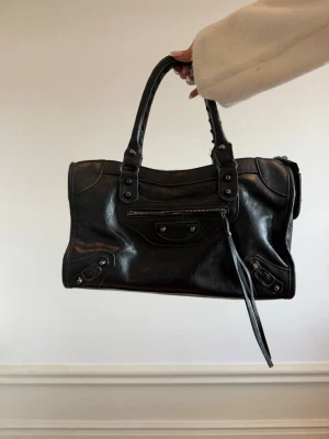 Svart Balenciaga City handväska - Svart Balenciaga City handväska i läder med klassiska nitar och dragkedjedetaljer. Väskan har dubbla handtag, dekorativ dragkedja framtill och en avtagbar läderrem. 