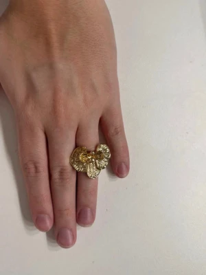 Guldfärgad ring med blomform - Unik guldfärgad ring i form av en blomma med detaljerad struktur och små pärlor i mitten. Ringen har en iögonfallande design som verkligen sticker ut och passar perfekt för dig som gillar statement-smycken. Materialet ser ut som metall med glansig finish. Vet inte exakta storleken men skulle gissa på M/L 