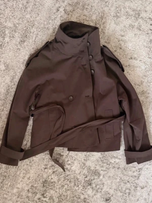 Bershka trenchcoat med bälte - Kort trenchcoat i mörkbrunt tyg med hög krage, dubbelknäppning och axelklaffar. Jackan har ett matchande bälte i midjan och uppvikta ärmslut. Klassisk och stilren modell som passar till många looks. Använd 1 gång! Som ny!!