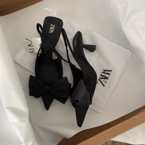 Svarta pumps med rosett från Zara - Säljer ett par svarta pumps från Zara med spetsig tå och stor dekorativ rosett framtill. Skorna har smal klack och öppen häl med justerbar rem. Materialet ser ut att vara en mix av textil och syntet. Perfekta för dig som vill ha en elegant och trendig look.