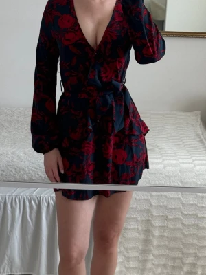 Blommig omlottklänning med volang - Svart klänning med röda rosor och omlottdetaljer. Väldigt feminin och smickrande modell med markerad midja och flowy ärmar. Perfekt till middag, fest eller dejt