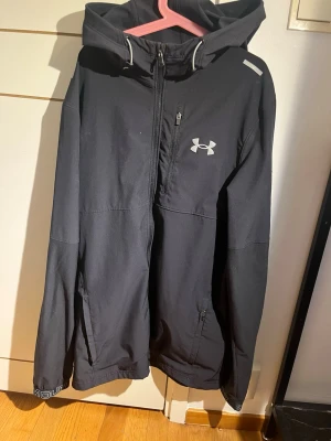 Svart vindjacka från Under Armour - Snygg svart vindjacka från Under Armour med huva och dragkedja framtill. Jackan har två sidofickor med dragkedja, en bröstficka och logga på bröstet. Tillverkad i ett lätt och slitstarkt material som passar perfekt för blåsiga dagar.