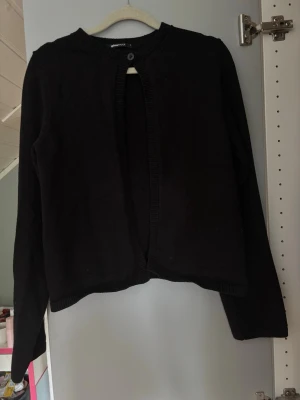 Svart kofta från Gina Tricot - Säljer en svart kofta från Gina Tricot med rund halsringning och en knapp upptill. Koftan har långa ärmar och ribbade kanter vid hals, ärmslut och nederkant. Enkel och stilren design som passar till det mesta.