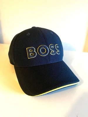 Marinblå BOSS keps med gul detalj - Snygg marinblå keps från BOSS med broderad logga i gult och blått framtill. Kepsen har böjd skärm med gul kant och är tillverkad i ett tåligt bomullstyg. Perfekt accessoar för en sportig och trendig look.