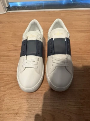 Valentino vita sneakers med blå detalj - Snygga vita sneakers från Valentino med bred mörkblå detalj över snörningen. Klassisk låg modell i skinn med vita snören och diskret logga på plösen. Perfekta för dig som vill ha en stilren och exklusiv look.