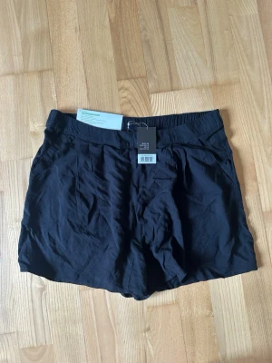 Svarta shorts med resårmidja - Snygga svarta shorts med resår i midjan och diskreta veck framtill. De har en avslappnad passform och är tillverkade i ett lätt och mjukt material som känns skönt mot huden. Perfekta för varma dagar och enkla att matcha med olika toppar.