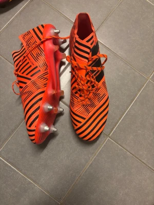     Adidas Nemeziz 17.1 SG - Säljer ett par grymma Adidas fotbollsskor i orange och svart med coolt mönster och snörning. Skorna har metall-dobbar för bästa grepp på planen och en strumpliknande passform för extra komfort. Perfekta för dig som vill sticka ut på matchen! Saknas 1 dobb annars i nyskick använd fåtal gånger