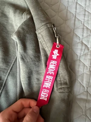 Grön bomberjacka från Alpha Industries - Säljer en klassisk grön bomberjacka från Alpha Industries med den ikoniska röda 'Remove Before Flight'-taggen på ärmen. Jackan har ficka med dragkedja på ärmen och ribbade muddar. Perfekt för dig som gillar streetwear och snygga detaljer. Passar för M.  Pris går att diskutera
