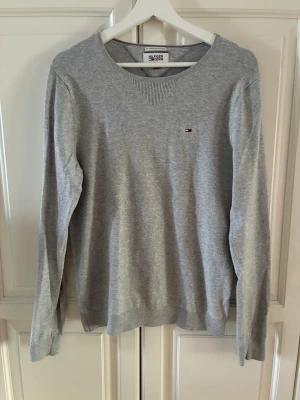 Grå tröja från Tommy Hilfiger - Klassisk grå tröja från Tommy Hilfiger med rund halsringning och diskret logga på bröstet. Tillverkad i mjukt bomullsmaterial och har ribbade muddar vid ärmslut och nederkant. Perfekt för en clean och stilren look.