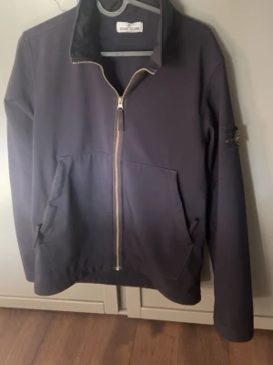 Svart softshelljacka från Stone Island - Snygg svart softshelljacka från Stone Island med dragkedja framtill och två stora fickor. Jackan har hög krage och den klassiska logotypen på ärmen. Perfekt för dig som gillar streetwear och vill ha en stilren look.