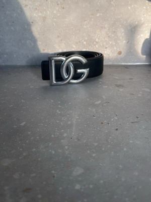 Svart DG bälte med silverdetalj - Snyggt svart bälte från Dolce & Gabbana med en stor, silverfärgad DG-logga som spänne. Bältet är stilrent och passar perfekt till jeans eller kostymbyxor. Klassisk design med exklusiv känsla. Pris kan diskuteras