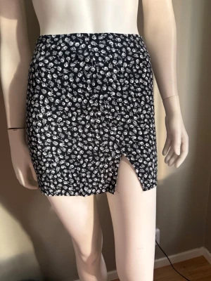 Svart blommig minikjol med slits strl XS  - Snygg svart minikjol med vita blommor och en liten slits framtill. Kjolen har en figurnära passform och är perfekt för dig som gillar trendiga detaljer. Tillverkad i ett mjukt och lätt material som känns skönt mot huden. Dold dragkedja baktill