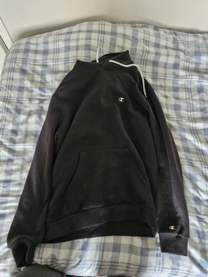 Svart hoodie från Champion - Svart hoodie från Champion med vit dragsko i huvan och klassisk Champion-logga på bröstet och ärmen. Stor känguruficka framtill och mjukt material som känns skönt mot huden. Perfekt för en chill och avslappnad stil.