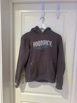 Grå hoodie från Hoodrich - Säljer en grå hoodie från Hoodrich med stor vit och turkos logga på bröstet och textdetaljer på ryggen. Hoodien har huva, känguruficka och ribbade muddar. Perfekt för dig som gillar streetwear och vill ha en chill look.(fläckarna är borta på denna hoodie)