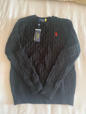 Svart kabelstickad tröja Polo Ralph Lauren - Kabelstickad svart tröja från Polo Ralph Lauren med klassisk röd broderad logga på bröstet. Tröjan har rund halsringning, långa ärmar och ribbade muddar. Perfekt för dig som gillar stilrena och tidlösa plagg. Mått: längd 60cm. Axelbredd 35cm.
