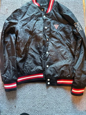 Svart bomberjacka från Guess - Snygg svart bomberjacka från Guess med vita och röda detaljer på krage, ärmslut och midja. Jackan har tryckknappar framtill, Guess-logga på bröstet och stor broderad text på ryggen. Tillverkad i glansigt syntetmaterial och har en klassisk college-look.