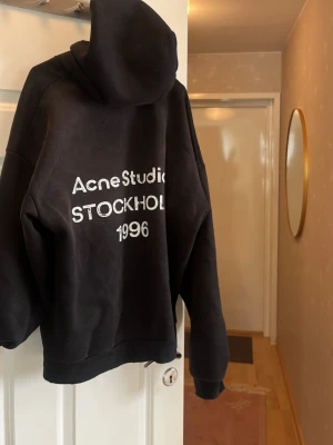 Svart hoodie Acne Studios Stockholm - Svart hoodie från Acne Studios med vit text 'Acne Studios STOCKHOLM 1996' på ryggen. Oversized passform med huva och ribbade muddar. Perfekt för en chill och trendig look. Materialet är mjukt och skönt bomullstyg.