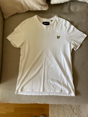 Vit t-shirt från Lyle & Scott - Vit t-shirt från Lyle & Scott med klassisk passform och rund hals. T-shirten har korta ärmar och en broderad gul logotyp på bröstet. Tillverkad i mjuk bomull som känns skön mot huden. Perfekt till jeans eller shorts.