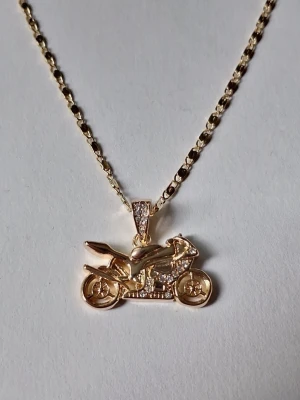 Guldfärgat halsband med motorcykelhänge - Unikt halsband i guldfärgat rostfritt stål med ett detaljerat motorcykelhänge. Hänget har små gnistrande stenar som ger extra bling och cool känsla. Kedjan är tunn och elegant, perfekt för dig som gillar statement-smycken med en sportig twist.