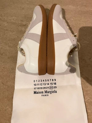 Maison Margiela  sneakers vit/beige - Maison Margiela sneakers i vitt läder med beige mockadetaljer och klassisk gummisula. Snygg retrostil med diskreta paneler och låg silhuett. Perfekta för dig som gillar minimalistisk design och exklusiva rial.