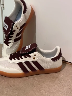 Adidas Wales Bonner sneakers vinröd/vit - Snygga Adidas Wales Bonner sneakers i samarbete med Wales Bonner. Skorna är vita med vinröda detaljer, klassiska tre ränder och grovstickade sömmar. Ovandel i mocka och läder, gummisula och bred plös med logga. Perfekta för dig som gillar unik stil.