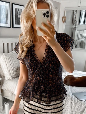 Svart blommig volangblus - Säljer en svart blus med små blommönster och volangdetaljer längs v-ringningen och nederkanten. Blusen är transparent och har korta, luftiga ärmar. Använd 1 gång, alltså nyskick! Helt slutsåld!!