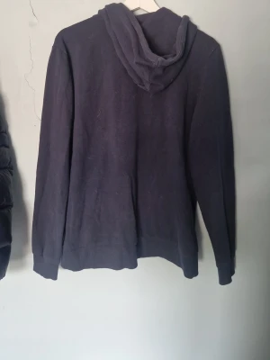 Mörkblå zip hoodie från H&M - Mörkblå hoodie från H&M med dragkedja framtill, två fickor och justerbar huva med snören. Tillverkad i mjukt bomullsmaterial och har ribbade muddar vid ärmslut och nederkant. Perfekt för en avslappnad och stilren look.