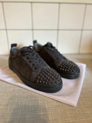Mörkgrå sneakers med nitar - Säljer ett par mörkgrå sneakers från Christian Louboutin i mocka med coola nitar på tån och klassisk röd sula. Skorna har svarta snören och en låg profil som ger en edgy och trendig look. Perfekta för dig som vill sticka ut med din stil.