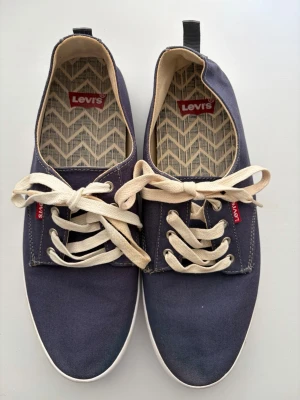 Blå Levi's canvas sneakers unisex - Snygga blå sneakers från Levi's i canvasmaterial med vita snören och vit sula. Skorna har klassisk låg modell och mönstrat innerfoder med Levi's-logga både på insidan och hälen. Perfekta för en avslappnad och stilren look.