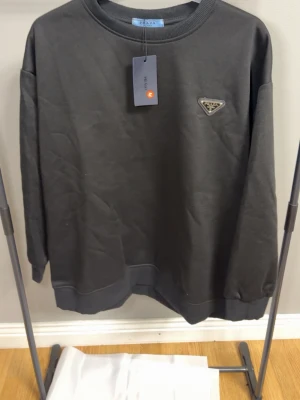 Svart sweatshirt från Prada - Snygg svart sweatshirt från Prada med klassisk rund halsringning och ribbade muddar. Tröjan har en ikonisk metallogo på bröstet och är tillverkad i mjukt material som känns skönt mot huden. Perfekt för dig som gillar stilrena och exklusiva plagg.